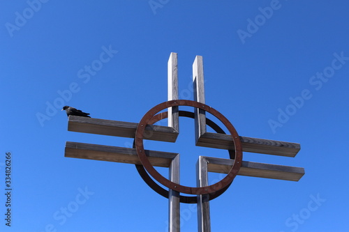 A black raven perching on a wooden cross on the top of a mountain isolated on clear blue sky.  photo