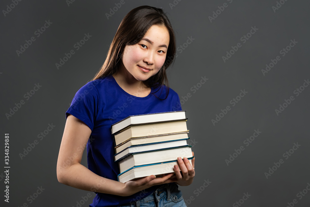 Asian student on grey background in studio holds stack of university books from library. Woman smiles, she is happy to graduate. Asian student on grey background in studio holds stack of university books from library. Woman smiles, she is happy to graduate.
