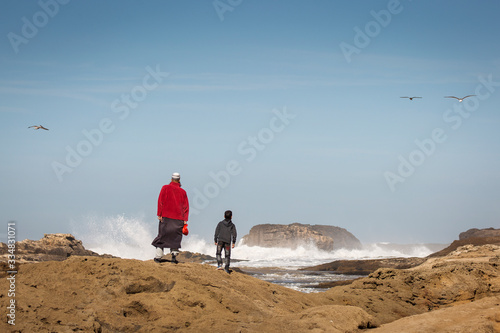 Father and son on rocks near to ocean with big waves and blue sky in background and seagulls flying around. Essaouira, Morocco