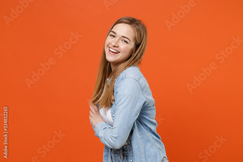 Side view of funny young woman girl in casual denim clothes posing isolated on orange background studio portrait. People lifestyle concept. Mock up copy space. Holding hands crossed, looking camera. © ViDi Studio
