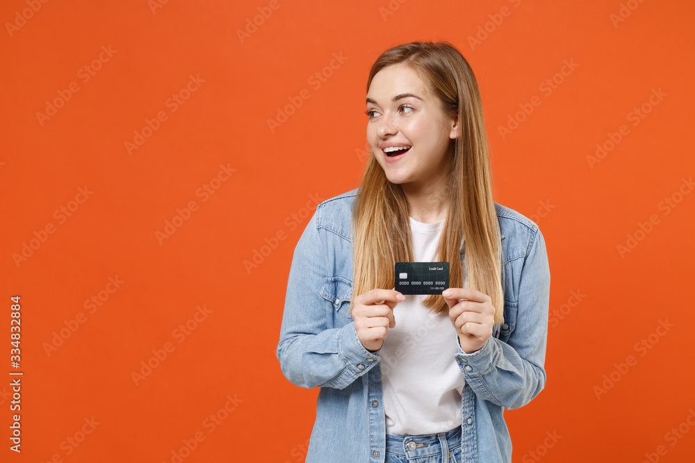 Cheerful young woman girl in casual denim clothes posing isolated on bright orange wall background studio portrait. People lifestyle concept. Mock up copy space. Hold credit bank card, looking aside. Cheerful young woman girl in casual denim clothes posing isolated on bright orange wall background studio portrait. People lifestyle concept. Mock up copy space. Hold credit bank card, looking aside.