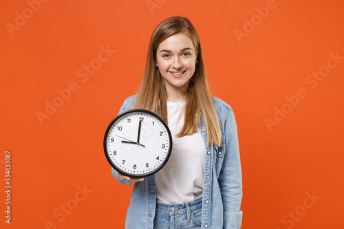 Smiling beautiful young woman girl in casual denim clothes isolated on bright orange wall background studio portrait. People sincere emotions lifestyle concept. Mock up copy space. Hold in hand clock.