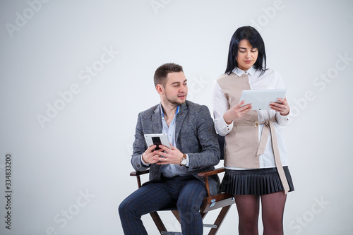 A successful man in a suit sits on a black chair and a woman stands next to look at the tablets. Future businessmen are smiling and talking about a new project.
