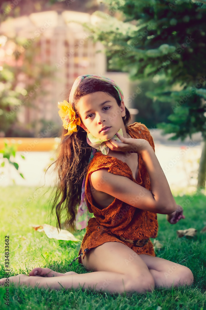 Beautiful little girl brunette with long hair and a hairpin in the form of a flower on her head sitting near a tree in the park. Summer vacation concept. Beautiful little girl brunette with long hair and a hairpin in the form of a flower on her head sitting near a tree in the park. Summer vacation concept.