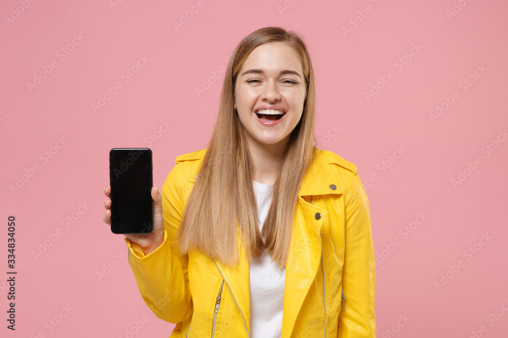 Fototapeta premium Laughing young woman girl in yellow leather jacket posing isolated on pastel pink background studio portrait. People lifestyle concept. Mock up copy space. Hold mobile phone with blank empty screen.