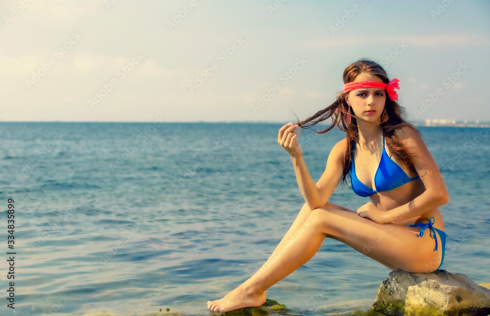 Travel Concept. Girl tourist sits on a mountain stone. Girl sitting on a cliff face over the sea. Clean sea and wild seascape.Young brunette girl with flower on her head enjoying vacation.Copy space