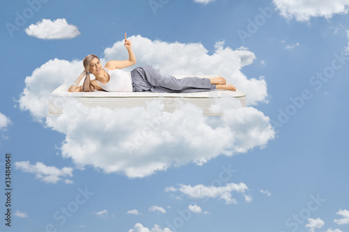 Young woman lying on a comfortbale mattress in pajamas, pointing up and floating on clouds photo