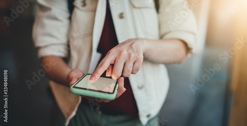 Frontal photo of young hipster girl in trendy white jeans jacket using modern smartphone typing sms message, closeup of female hands using cellphone with blank screen indoor, flare light effect