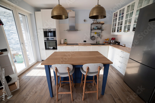 White, bright kitchen with blue island and double brass pendant lights. Central view.