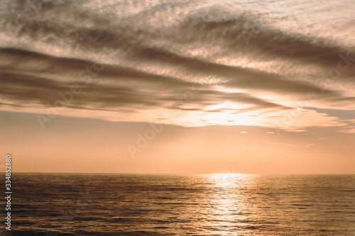 Fototapeta Naklejka Na Ścianę i Meble -  Beige sunset over the sea. The sun comes out from behind a undulating cloud over a quiet surface of the water