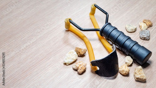 Man hand take stone with slingshot small hand-powered projectile weapon on wooden background. Classic form Y-shaped frame held in off hand with two rubber strips. Pocket that holds projectile photo