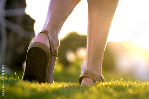 Close up of woman feet in summer sandals shoes walking on spring lawn covered with fresh green grass.