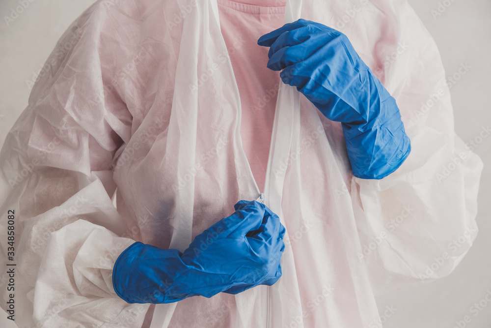 Fototapeta premium Doctor epidemiologist puts on white protective overalls, fastens a zipper, hands close-up. Hands in protective rubber gloves. Personal protective equipment concept.
