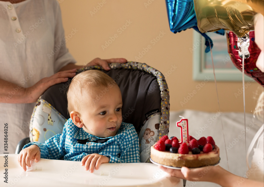Naklejka premium One year old cute boy and berries cake with a candle. Boy wearing celebration shirt, sitting on a highchair. First Happy Birthday at home. Family concept.