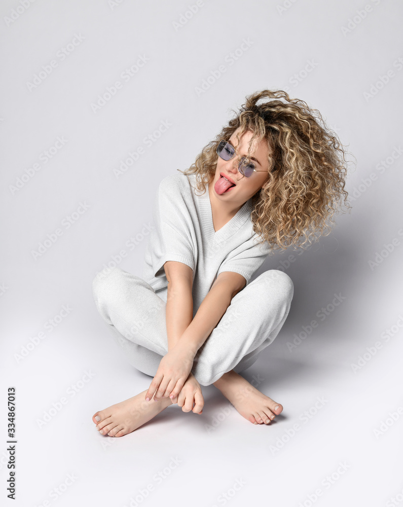 Blonde curly model in blue sunglasses, white suit of pants and blouse, barefoot. Showing tongue sitting on floor isolated on white Blonde curly model in blue sunglasses, white suit of pants and blouse, barefoot. Showing tongue sitting on floor isolated on white