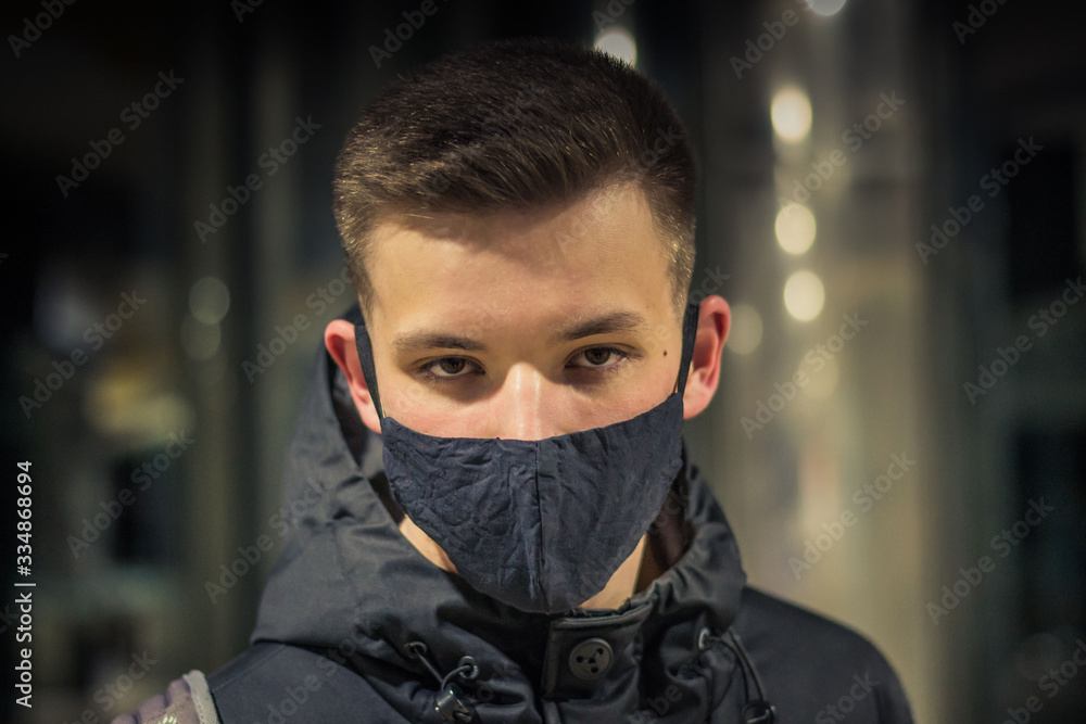 Closeup portrait of a young guy in a medical mask. Pandemic. Coronavirus. Quarantine. Covid19. Student in a protective mask. Gloomy young man in a black mask and black clothes on a dark background. Closeup portrait of a young guy in a medical mask. Pandemic. Coronavirus. Quarantine. Covid19. Student in a protective mask. Gloomy young man in a black mask and black clothes on a dark background.