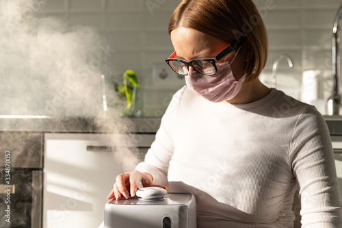 Woman in face protective mask holding in her hands working aroma oil humidifier in sunny room. Humidification of air in the apartment during the period of self-isolation due to coronavirus pandemic photo