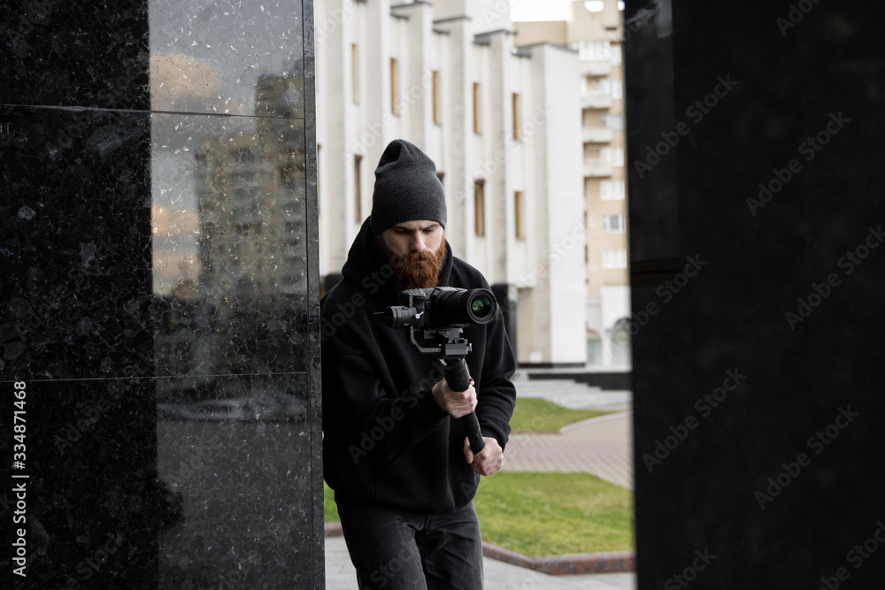 Bearded Professional videographer in black hoodie holding professional camera on 3-axis gimbal stabilizer. Filmmaker making a great video with a professional cinema camera. Cinematographer. Bearded Professional videographer in black hoodie holding professional camera on 3-axis gimbal stabilizer. Filmmaker making a great video with a professional cinema camera. Cinematographer.