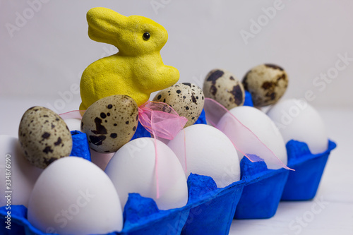 white chicken and quail eggs in a blue uniform, a tray and a yellow easter bunny with a pink bow on a light wooden background