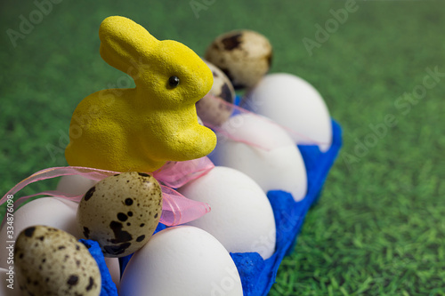 white chicken and quail eggs in a blue uniform, a tray and a yellow Easter bunny with a pink bow on a background of green grass