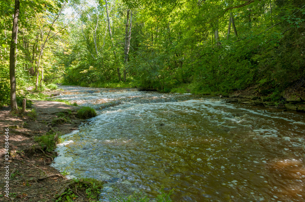 Wide view of small Baird Creek waterfall and rapids, flowing over the Niagara Escarpment at Baird Creek, Baird Creek Parkway, Green Bay, WI. Spring, summer rushing water. Wide view of small Baird Creek waterfall and rapids, flowing over the Niagara Escarpment at Baird Creek, Baird Creek Parkway, Green Bay, WI. Spring, summer rushing water.