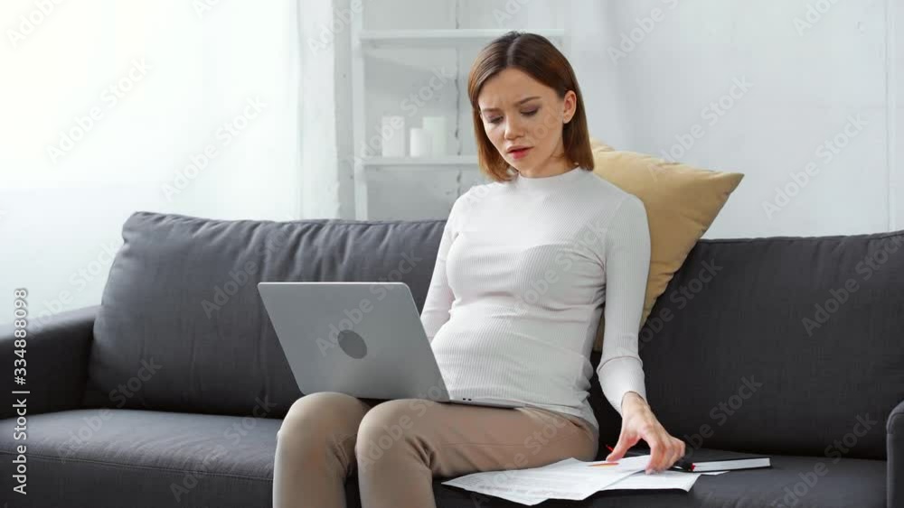 Worried pregnant freelancer with laptop and documents on couch Worried pregnant freelancer with laptop and documents on couch