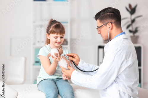 Pediatrician with toy showing little girl how to use stethoscope in clinic