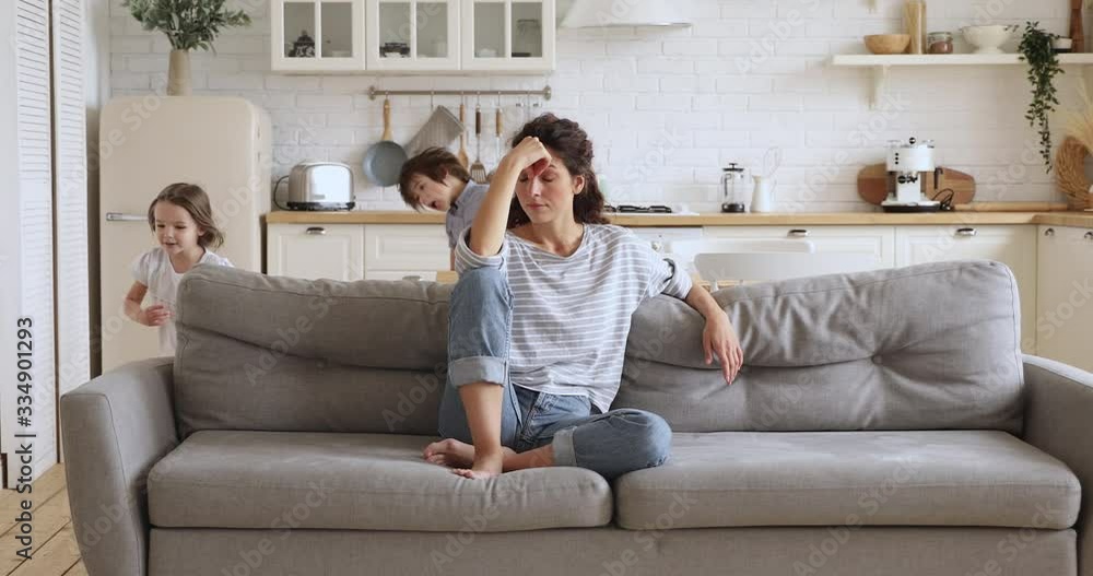 Tired upset young mother sit on sofa feel frustrated about active noisy kids running playing in kitchen, stressed exhausted fatigued single mom annoyed disturbed about disobedient difficult children
