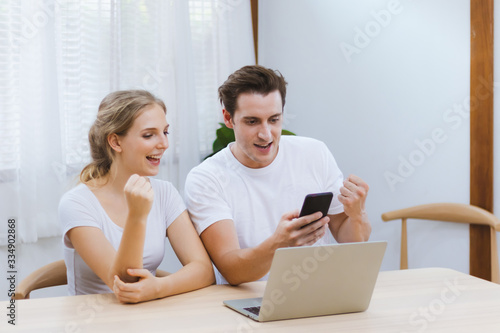 Smiling caucasian couple celebrating together for success of entrepreneur business online or job achievement or education exam result via smartphone at home. Internet & technology photo concept