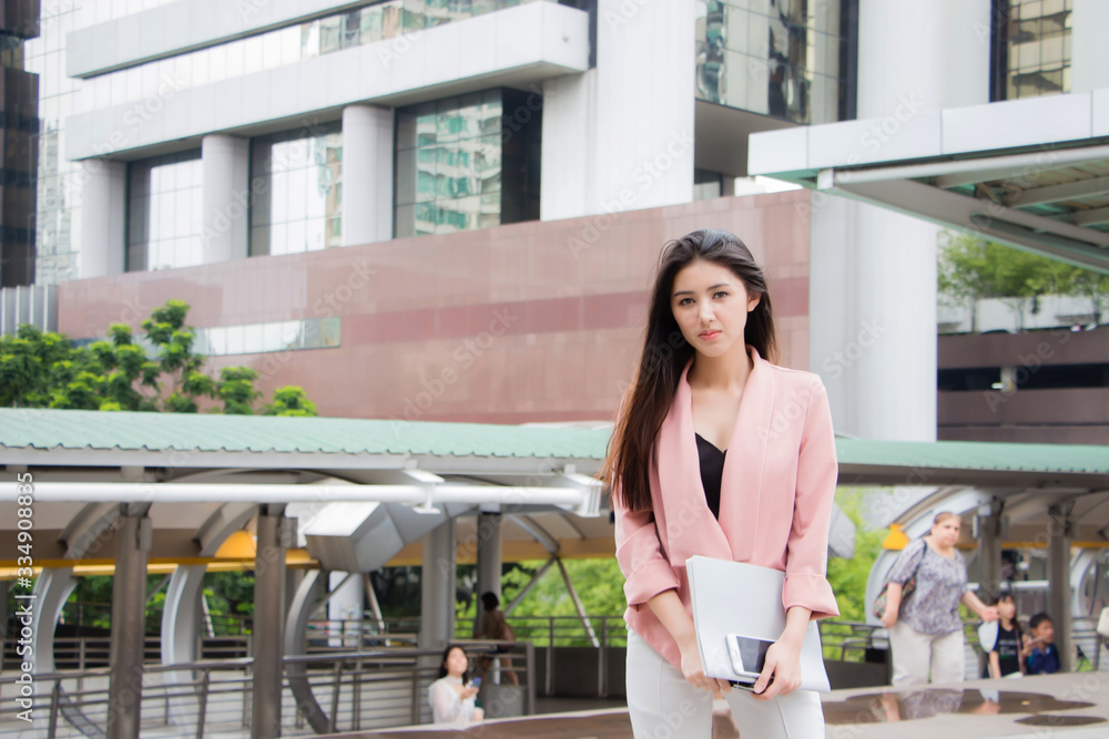 A beautiful Asian woman, Thai, is wearing pink work clothes and white pants standing and holding documents and mobile phones in front of a tall building in the metropolis. She looked at the camera