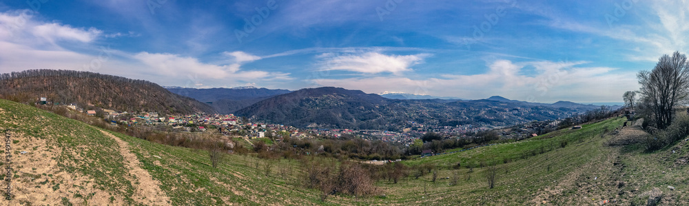 View of the outskirts of the city from above in sunny weather. Houses and mountains under a blue cloudy sky. Panoramic image of the Caucasus mountains.
