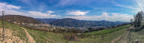 View of the outskirts of the city from above in sunny weather. Houses and mountains under a blue cloudy sky. Panoramic image of the Caucasus mountains.