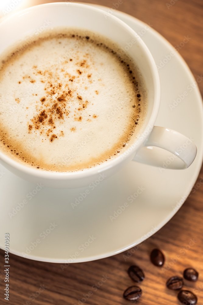Eine Kaffeetasse mit gerösteten Kaffeebohnen auf einem hölzernenen Tisch. Sonneneinstrahlung, Reflexionen. Ein Cappuccino mit Milchschaum und gemahlenem Kaffee. Serviert in einem Café. Milchkaffee Eine Kaffeetasse mit gerösteten Kaffeebohnen auf einem hölzernenen Tisch. Sonneneinstrahlung, Reflexionen. Ein Cappuccino mit Milchschaum und gemahlenem Kaffee. Serviert in einem Café. Milchkaffee