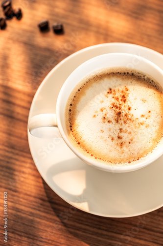 Eine Kaffeetasse mit gerösteten Kaffeebohnen auf einem hölzernenen Tisch. Sonneneinstrahlung, Reflexionen. Ein Cappuccino mit Milchschaum und gemahlenem Kaffee. Serviert in einem Café. Milchkaffee