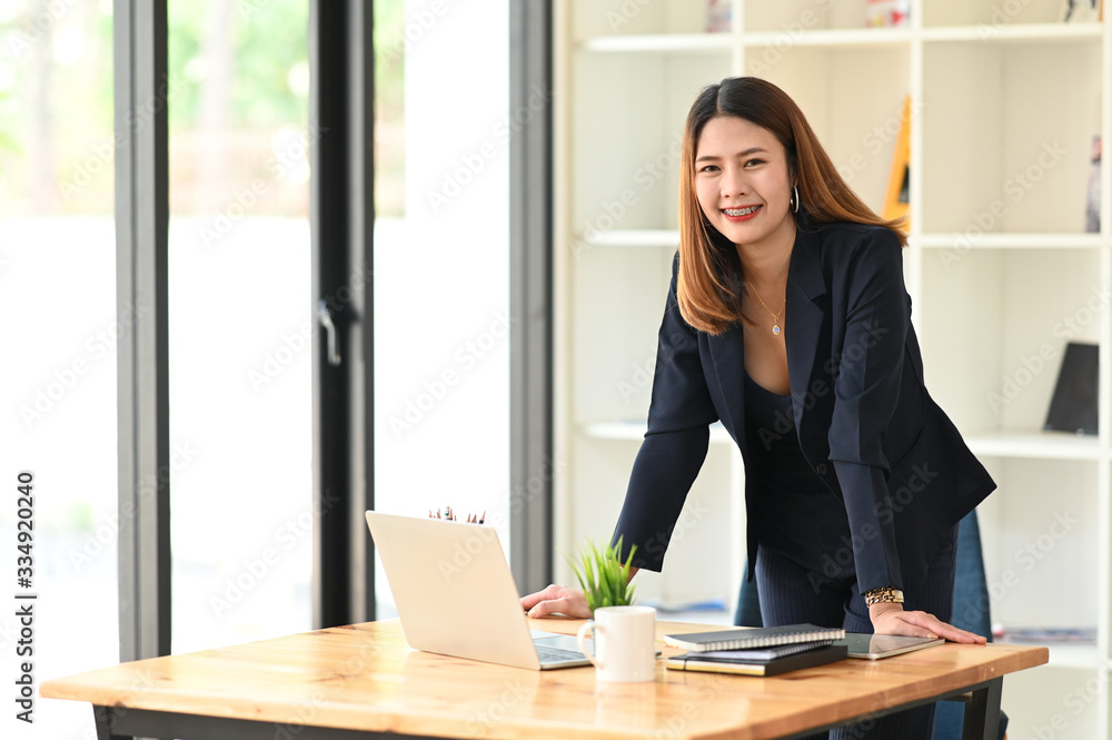 Photo of beautiful businesswoman standing up at her wooden working desk over the comfortable workplace as background. Comfortable work-space/Working at home concept. Photo of beautiful businesswoman standing up at her wooden working desk over the comfortable workplace as background. Comfortable work-space/Working at home concept.