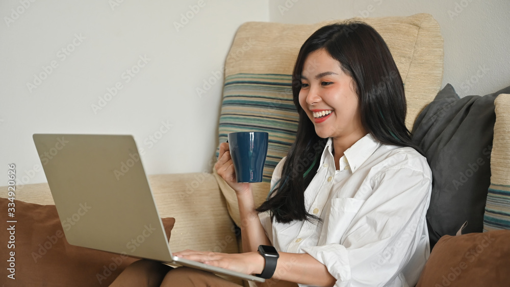 Young beautiful woman relaxing on her holiday/using a computer laptop and enjoying with her coffee while sitting at the comfortable couch over orderly living room as background. Young beautiful woman relaxing on her holiday/using a computer laptop and enjoying with her coffee while sitting at the comfortable couch over orderly living room as background.