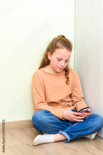 Girl sitting on floor and watching webinar on the smart phone in a home with a wall on background.