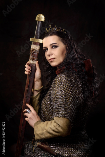 A dark-haired woman with a sword and crown  a Scandinavian warrior in a chain mail against a dark background