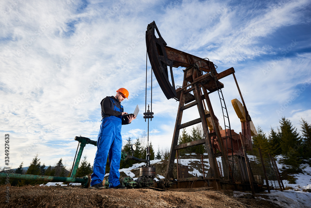 Oil worker using laptop, controlling work of petroleum pump jack. Drill operator in work overalls and helmet near oil pumping unit under cloudy sky. Concept of petroleum industry and oil extraction. Oil worker using laptop, controlling work of petroleum pump jack. Drill operator in work overalls and helmet near oil pumping unit under cloudy sky. Concept of petroleum industry and oil extraction.