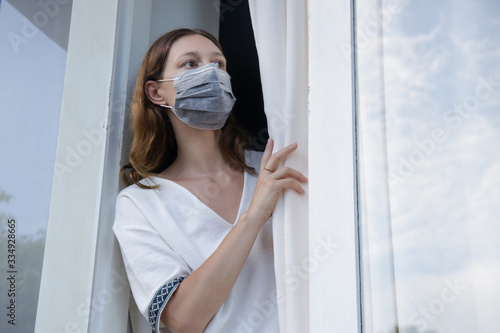 Young woman with surgical disposable mask looking outside through window. Bored and completely alone at home. Coronavirus outbreak and self-quarantining concept.