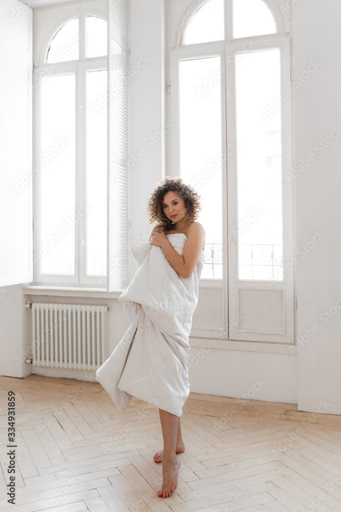 Beautiful sweet cheerful tender happy natural sleepy girl with Afro curls wrapped in a blanket wakes up and enjoys the morning looks out the window against the background of large vintage Windows Beautiful sweet cheerful tender happy natural sleepy girl with Afro curls wrapped in a blanket wakes up and enjoys the morning looks out the window against the background of large vintage Windows