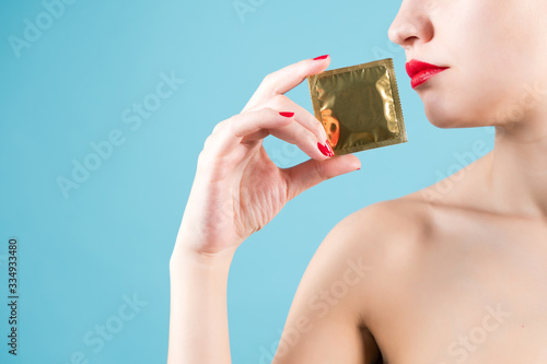 A young girl holds a condom near red lips with her fingers with red nail polish.