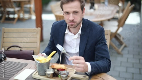 young smiling businessman is eating his hamburger from his plate of  French fries, sauces, sitting at a table with his briefcase next to him at a terrace