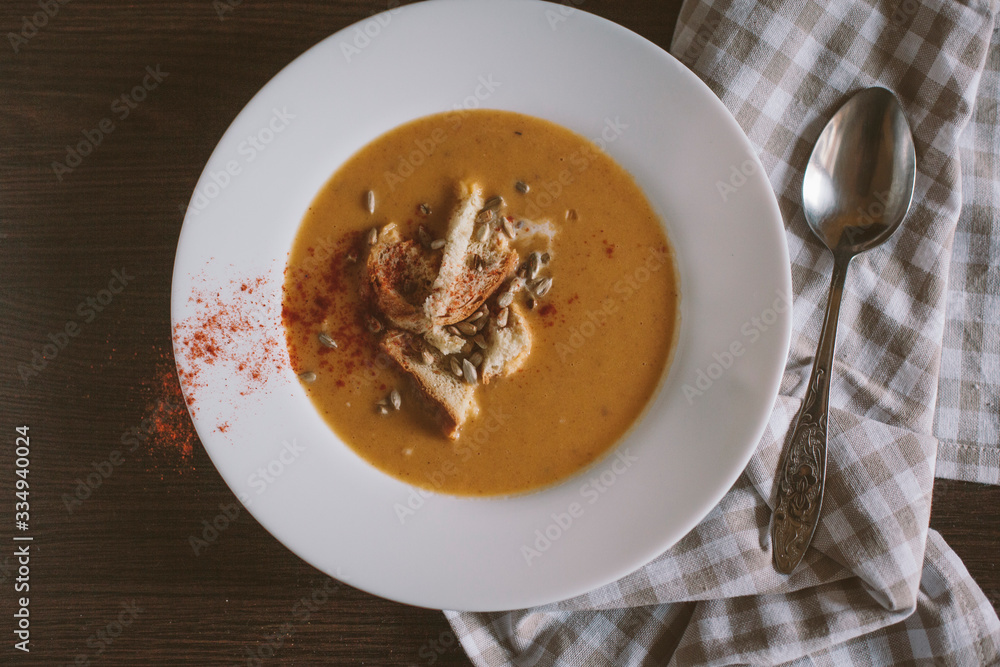 Pumpkin puree soup with crackers and seeds sprinkled with chili peppers in a large plate on a dark wooden background. Next to a spoon and a linen towel. Healthy and wholesome food. Pumpkin puree soup with crackers and seeds sprinkled with chili peppers in a large plate on a dark wooden background. Next to a spoon and a linen towel. Healthy and wholesome food.