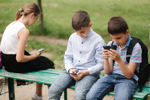 Two boys and girl use their phones during school breack. Cute boys sitting on the bench and play online games photo