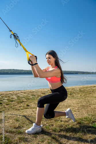 Young slim woman doing suspension training with fitness straps outdoors near the lake at daytime. Healthy lifestyle.