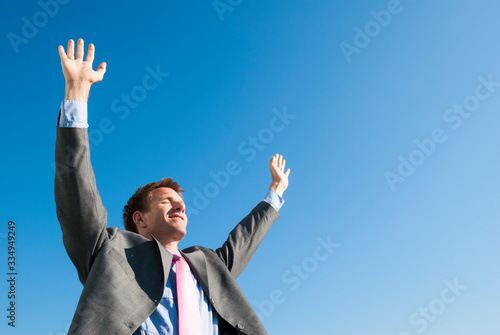 Smiling businessman standing outdoors holding arms up facing the sun against clear blue sky
