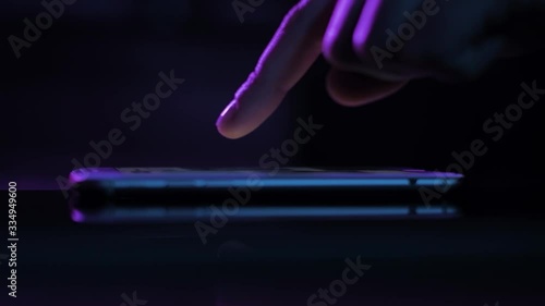 Young woman browsing internet on smart phone at night; girls finger tapping keypad, scrolling touchscreen of mobile device, purple blue low light; extreme close up side view