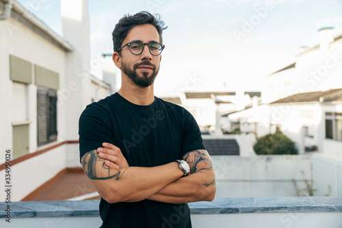 Confident stylish guy with tattoos posing on apartment balcony or terrace. Young man in glasses standing outside with arms crossed and looking at camera. Male portrait concept