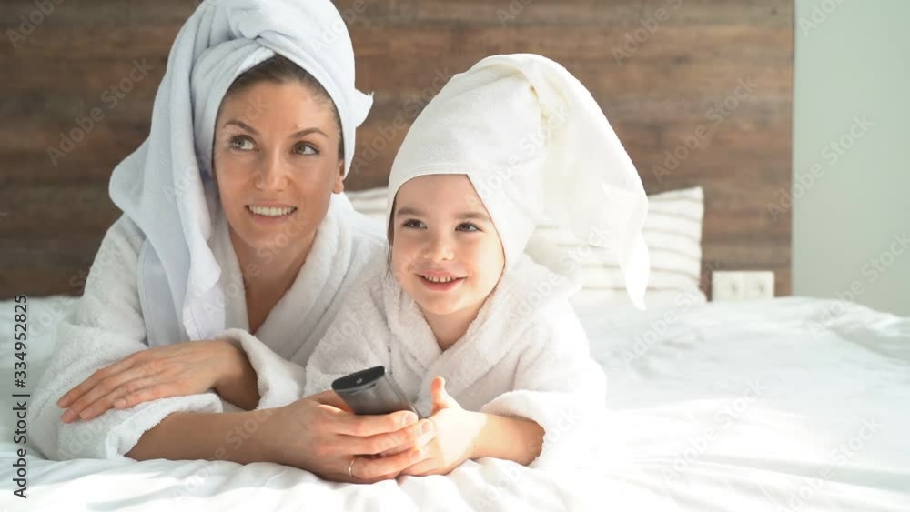 Beautiful mother with a little daughter watching TV lying on the bed in bathrobes and towels. Quarantine and self-isolation at home.